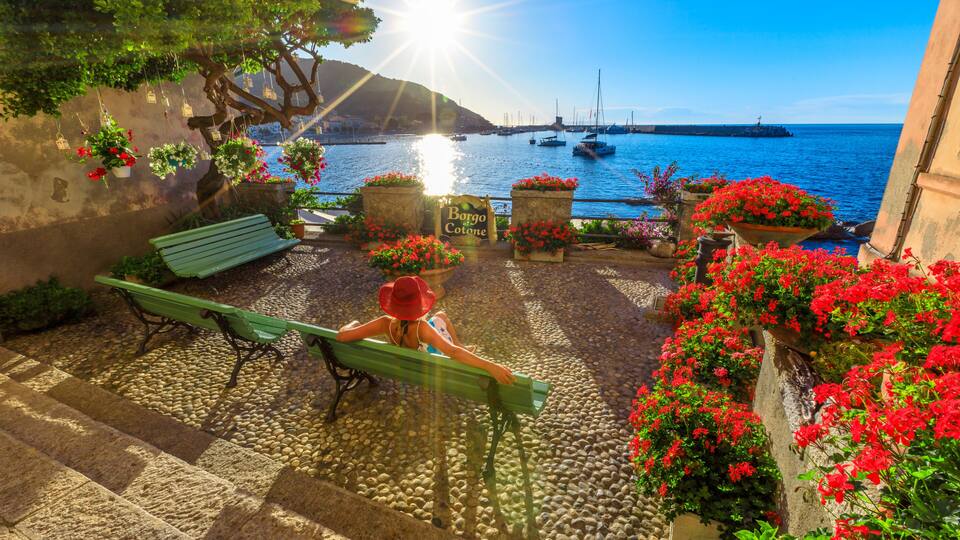 A coastal scene with a bench, hanging flower pots, and a sign reading 'Borgo Cotone.'