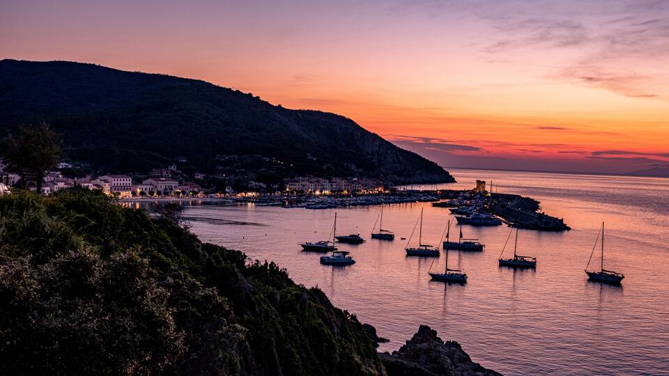 A coastal town at sunset with boats docked along the pier, a mountain in the background, and buildings lining the shore.