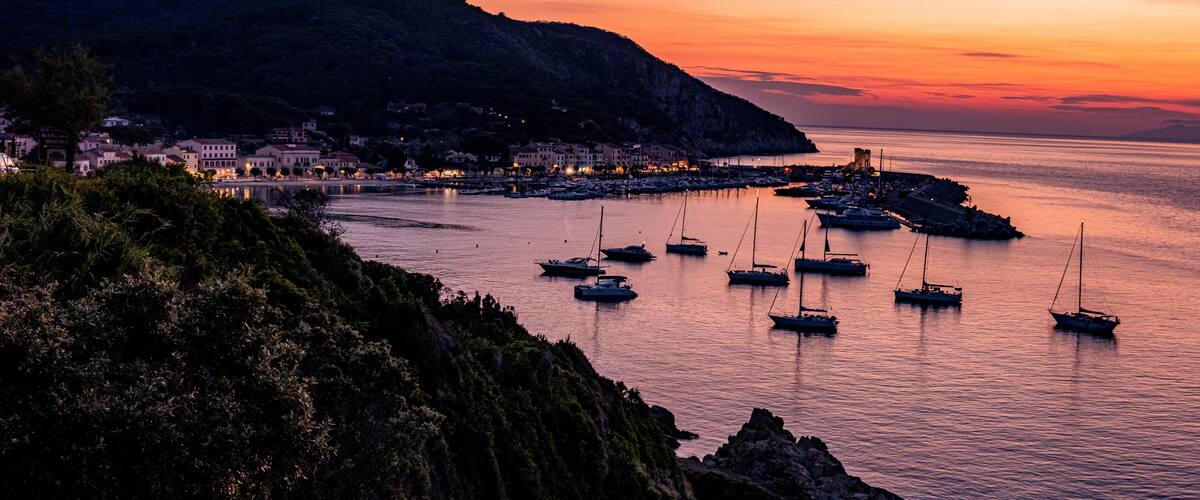 A coastal town at sunset with boats docked along the pier, a mountain in the background, and buildings lining the shore.