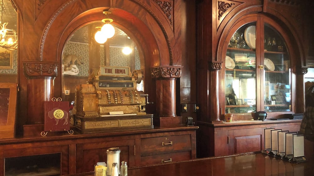 An ornate wooden bar with a vintage cash register labeled "The Irma" and a cash drawer.