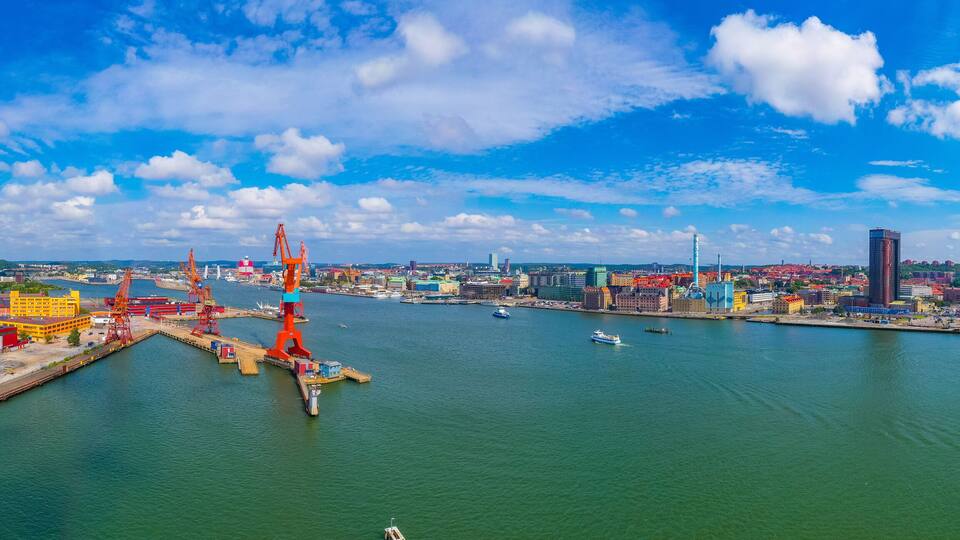 A harbor with cranes, a large ship, and a cityscape with buildings and a clear blue sky.