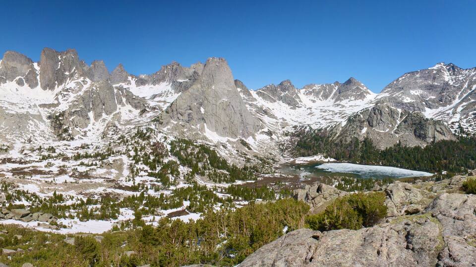 A rugged mountain landscape with snow-capped peaks, a glacier, and dense forested areas.