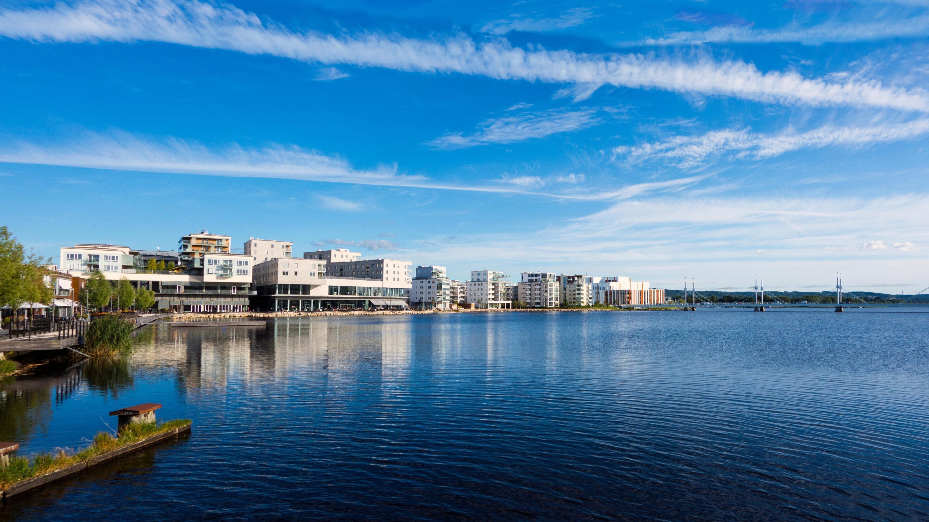 A waterfront cityscape with modern buildings, a calm river, and a bridge in the distance.