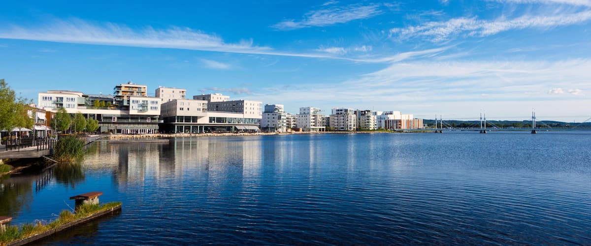 A waterfront cityscape with modern buildings, a calm river, and a bridge in the distance.