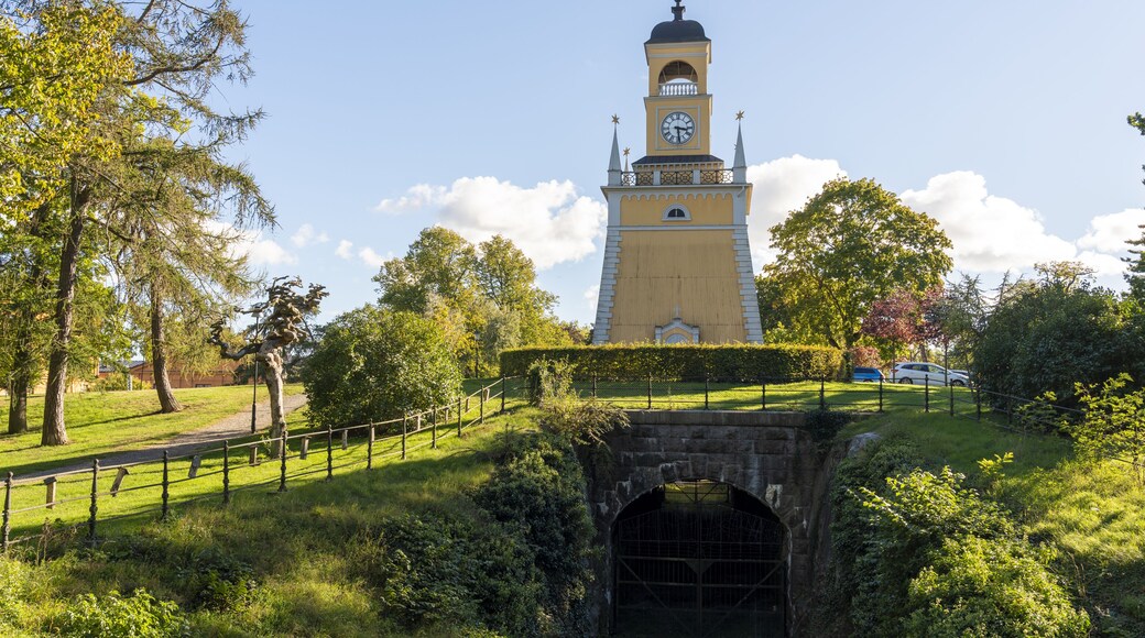 View of Amiralitetsklockstapeln clock tower standing tall above a moss-covered stone tunnel, framed by vibrant green foliage under blue sky, Karlskrona, Blekinge County, Sweden.