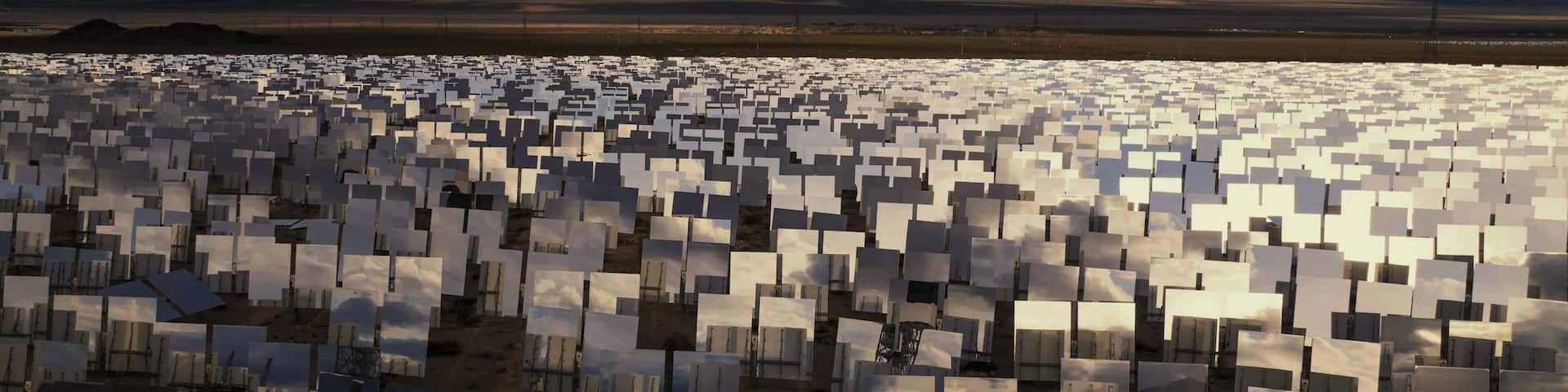 Aerial view of a vast expanse of solar mirrors glinting under the desert sun against a backdrop of distant mountains and dramatic clouds, California, San Bernardino County, United States.