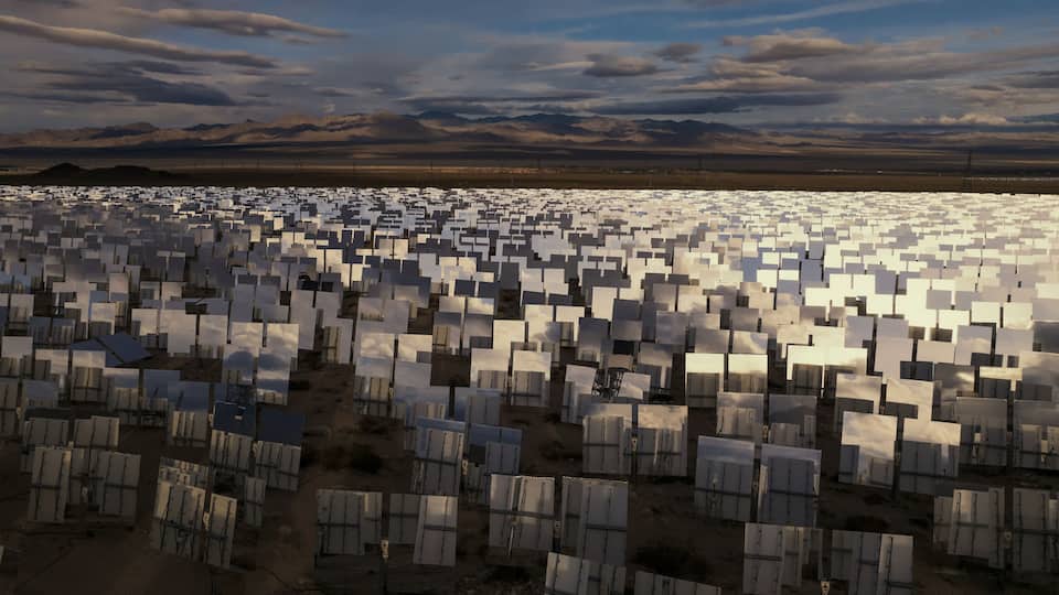 Aerial view of a vast expanse of solar mirrors glinting under the desert sun against a backdrop of distant mountains and dramatic clouds, California, San Bernardino County, United States.
