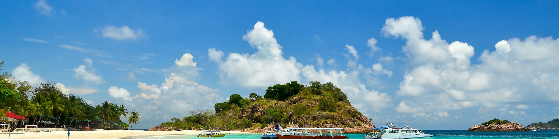 Uma praia com águas turquesa cristalinas, uma pequena ilha com vegetação densa e alguns barcos.