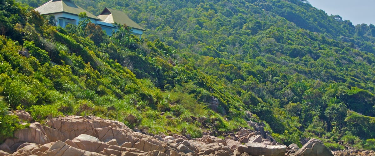 A beach with large rocks, a white sandy shore, and a mountain covered in dense green foliage in the background.