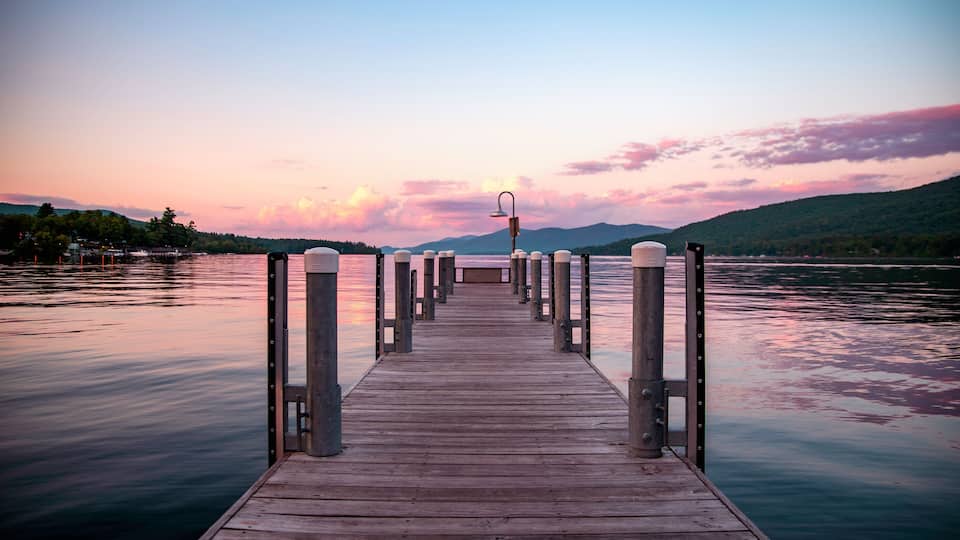 A wooden pier extending into calm waters, surrounded by hills and a sunset sky.