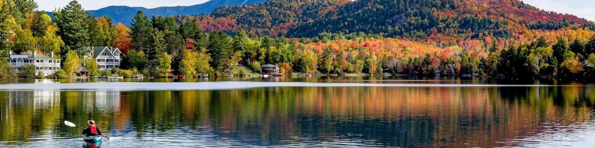 A calm lake with a kayaker, autumn-colored trees, and a mountain in the background.