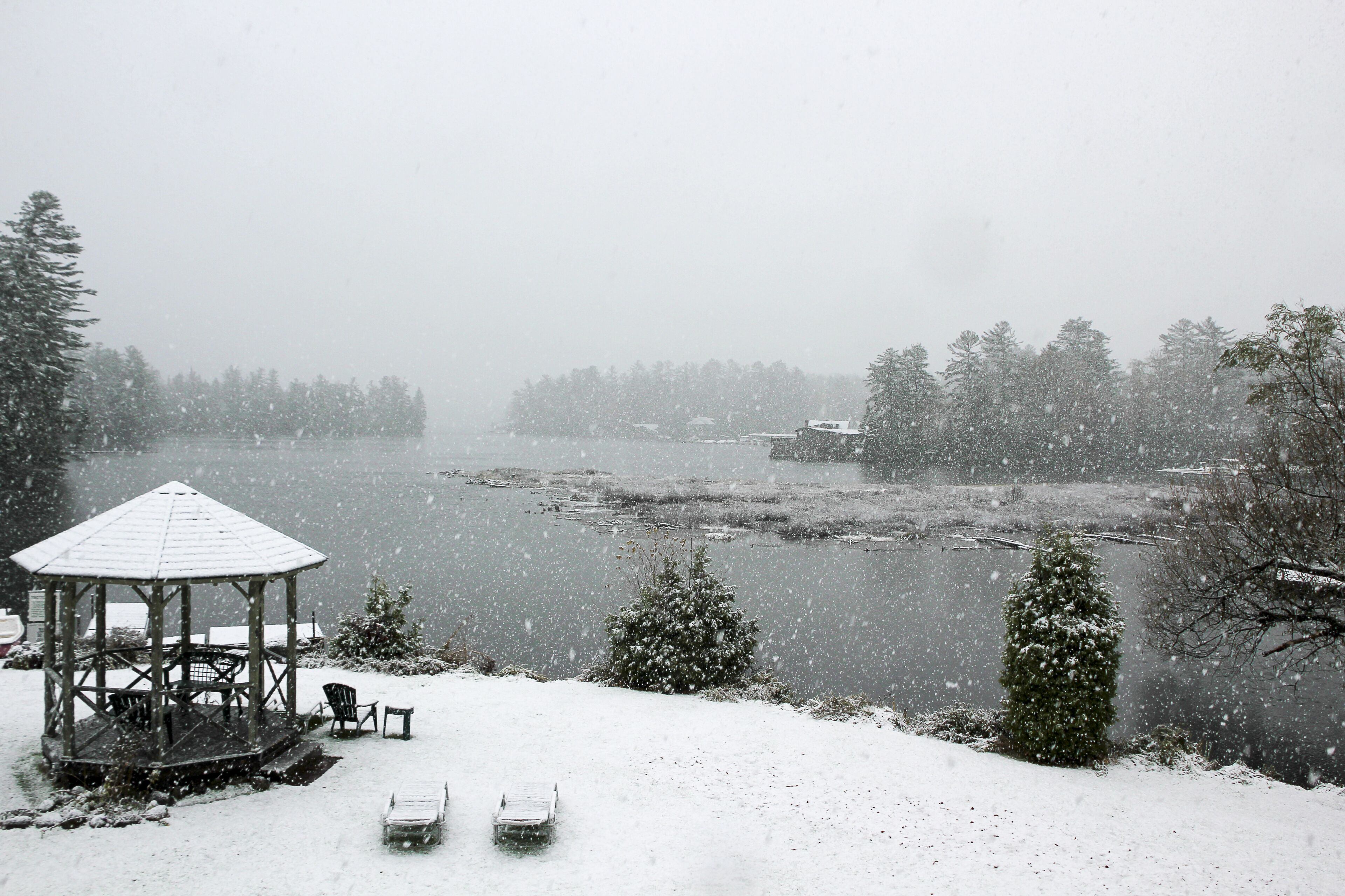 A gazebo, snow-covered chairs, a snow-laden tree, and a lake.