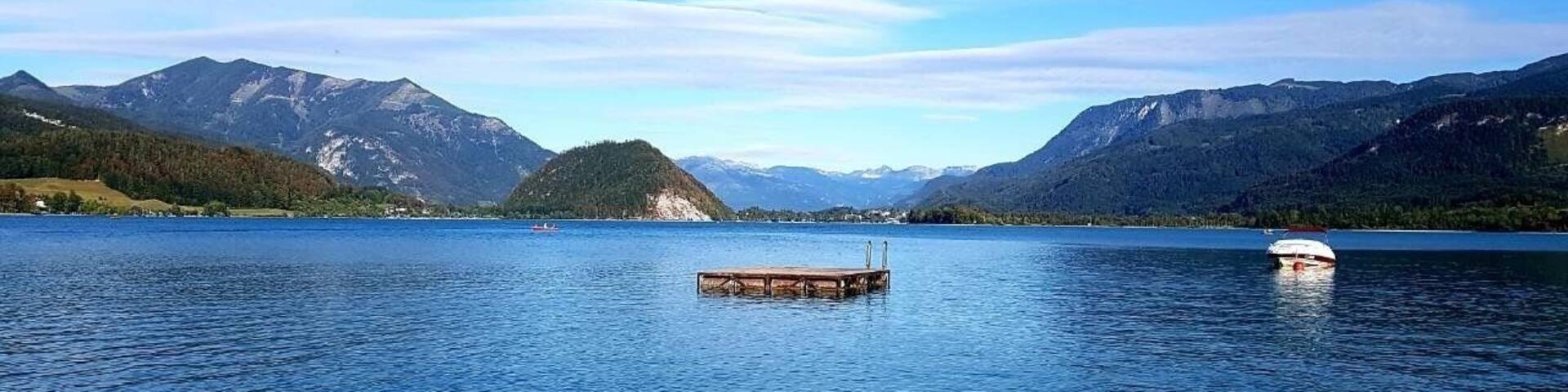 A calm lake with a wooden dock and a small boat, surrounded by mountains and clear skies.