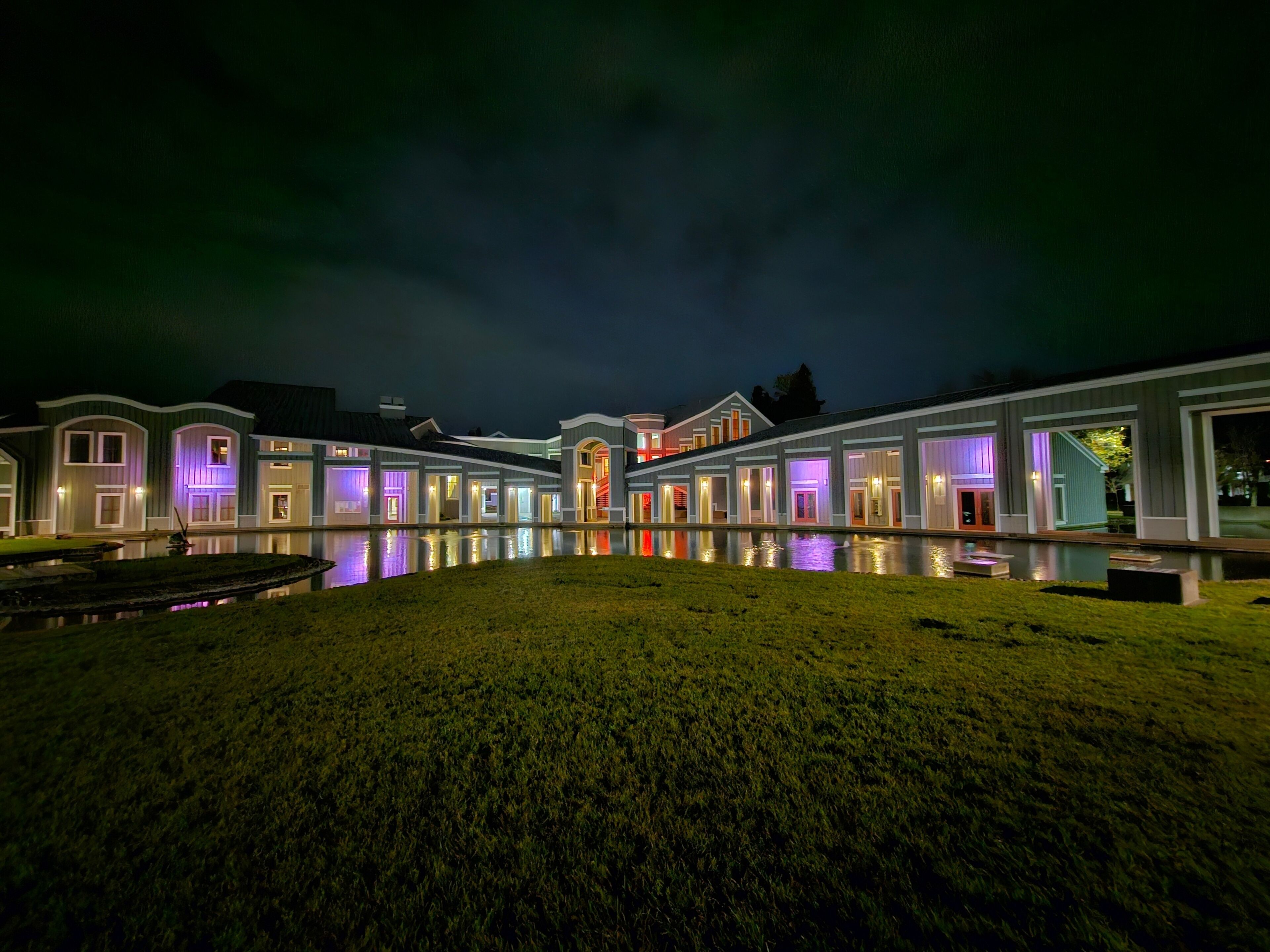 Un gran complejo de edificios iluminado de noche, con un elemento acuático reflectante al frente.