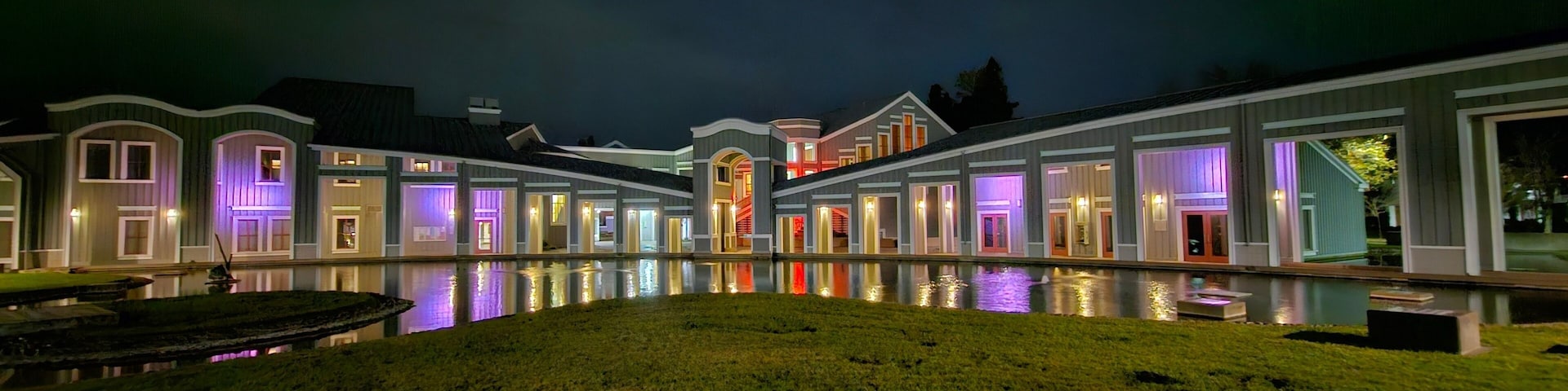 Un gran complejo de edificios iluminado de noche, con un elemento acuático reflectante al frente.