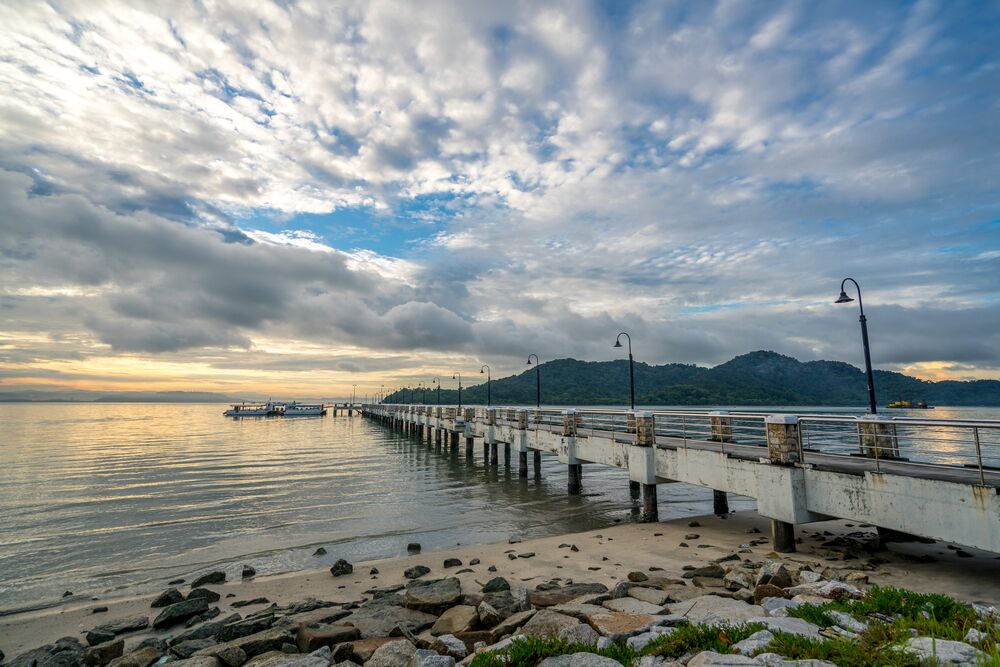 A pier extending into calm waters with a mountainous backdrop under a cloudy sky.