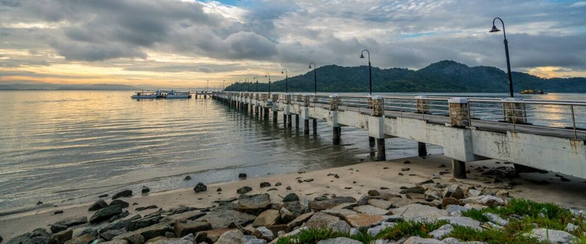 A pier extending into calm waters with a mountainous backdrop under a cloudy sky.