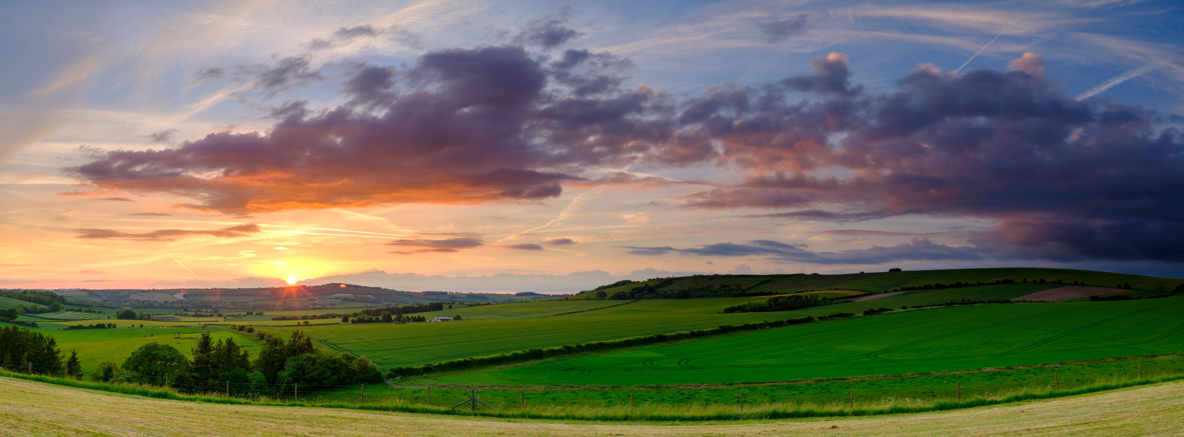 A landscape with rolling green hills, a clear sky with scattered clouds, and the sun setting or rising.