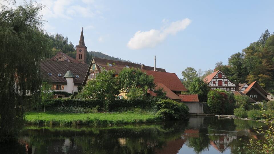 A riverside town with traditional half-timbered houses, a church with a tall steeple, and a calm water body reflecting the buildings.