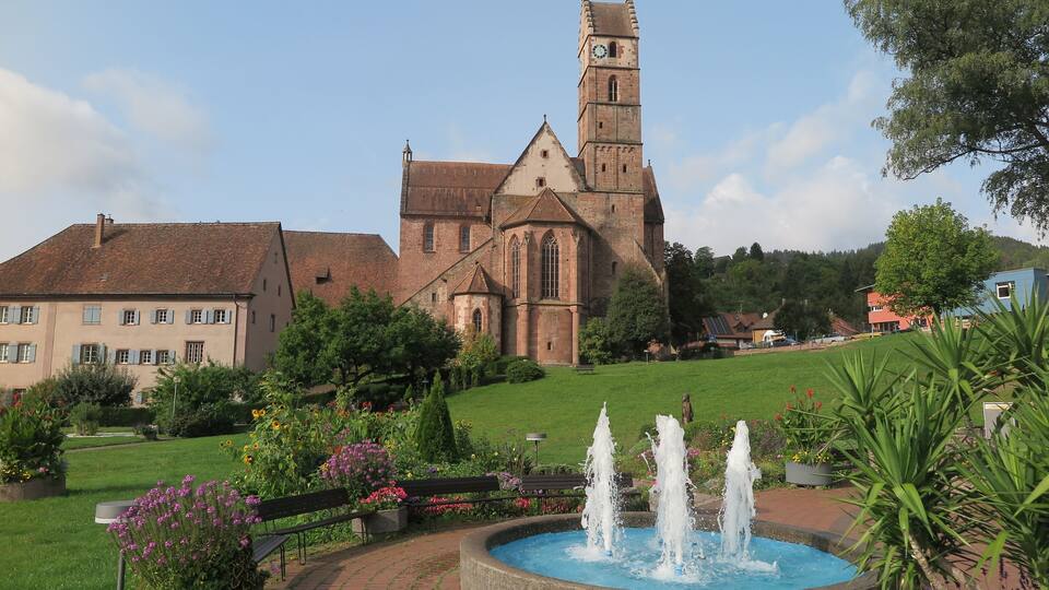 A church with a clock tower, a fountain, and surrounding greenery.