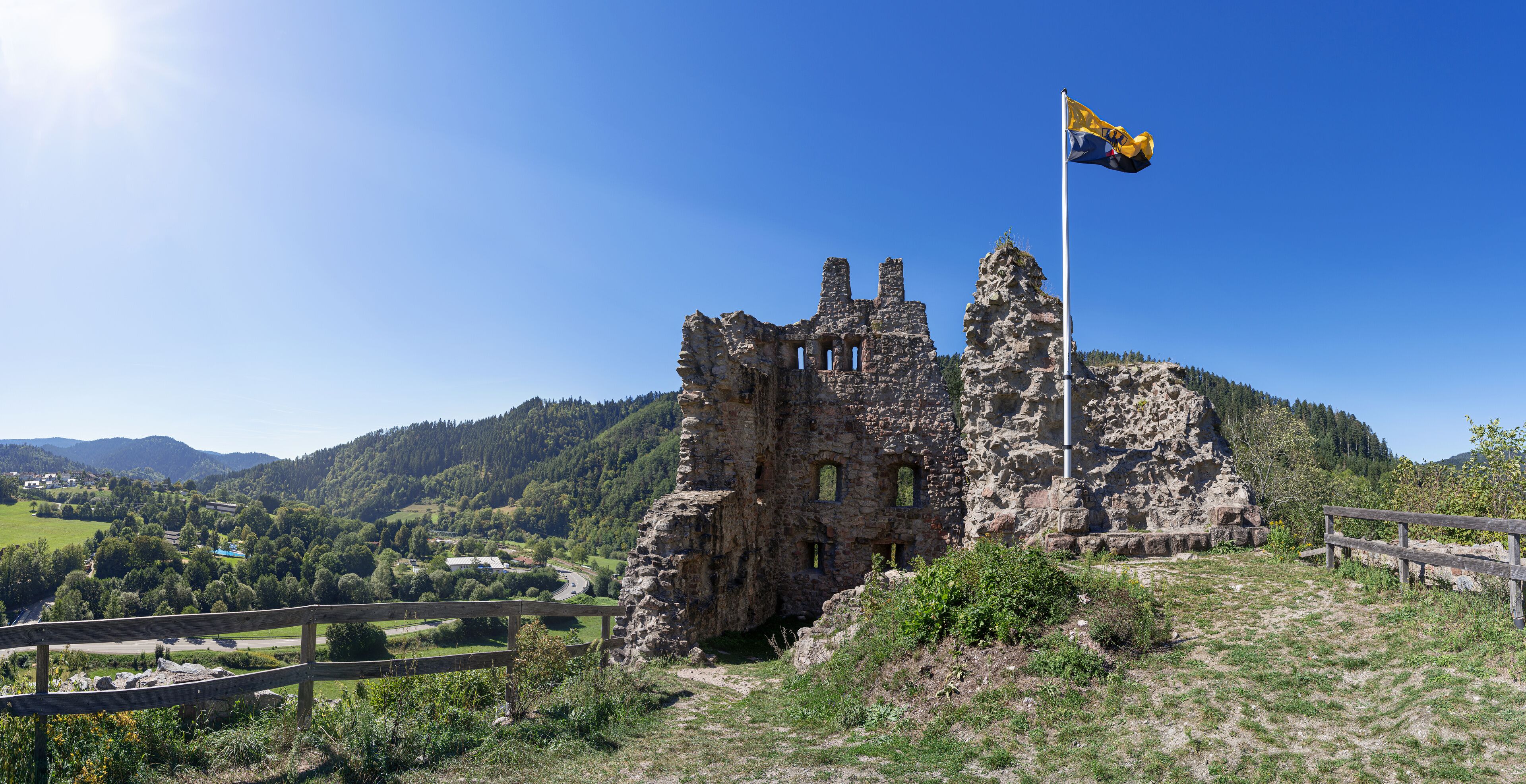 A historic stone castle with a flagpole, surrounded by a wooden fence and lush greenery.