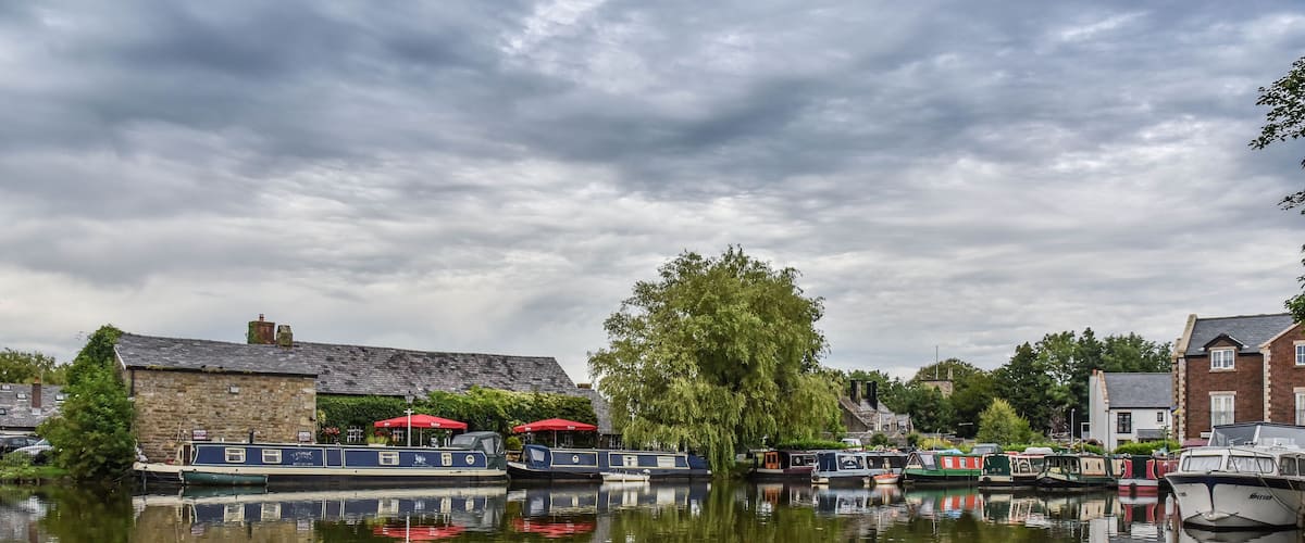 A calm river with boats docked, a stone building, and a large tree reflecting in the water.