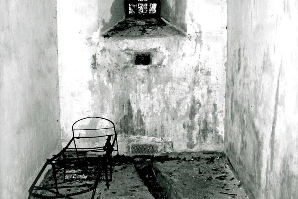 Una habitación estrecha y tenuemente iluminada, con una ventana con barrotes, una silla de metal y suelo de hormigón.
