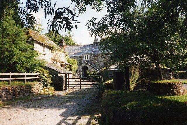 A rural stone house with a wooden gate, surrounded by trees and a gravel path.