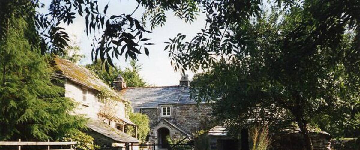 A rural stone house with a wooden gate, surrounded by trees and a gravel path.