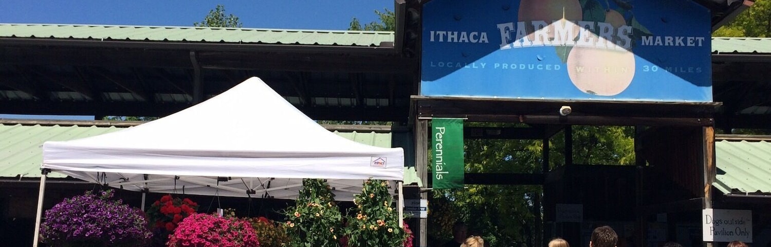 Un mercado de agricultores con un letrero azul que dice "ITHACA FARMERS MARKET" y una carpa blanca que vende flores.