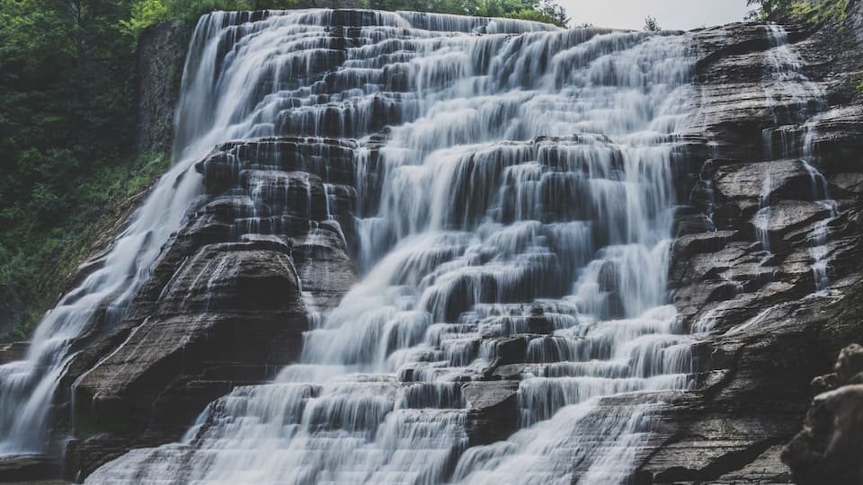 A multi-tiered waterfall cascading over dark rock formations surrounded by lush greenery.