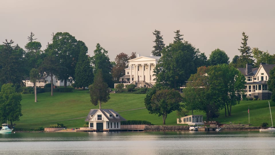 A lake with boats, a white house with columns, and a smaller building on a grassy hill.