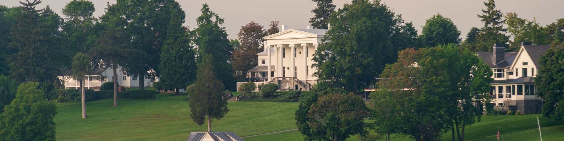 Un lago con barcos, una casa blanca con columnas y una edificación más pequeña en una colina cubierta de hierba.