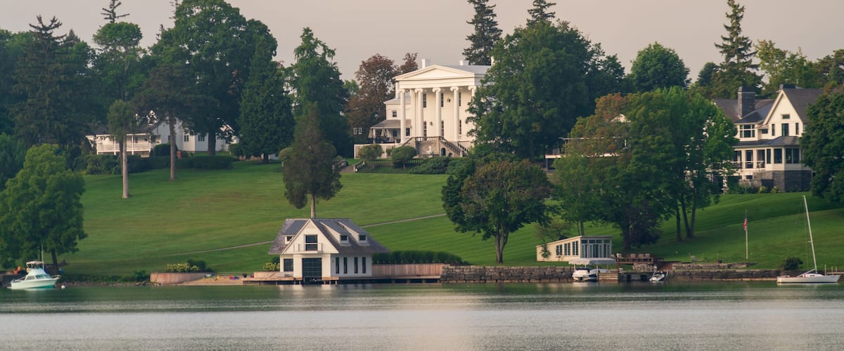 A lake with boats, a white house with columns, and a smaller building on a grassy hill.
