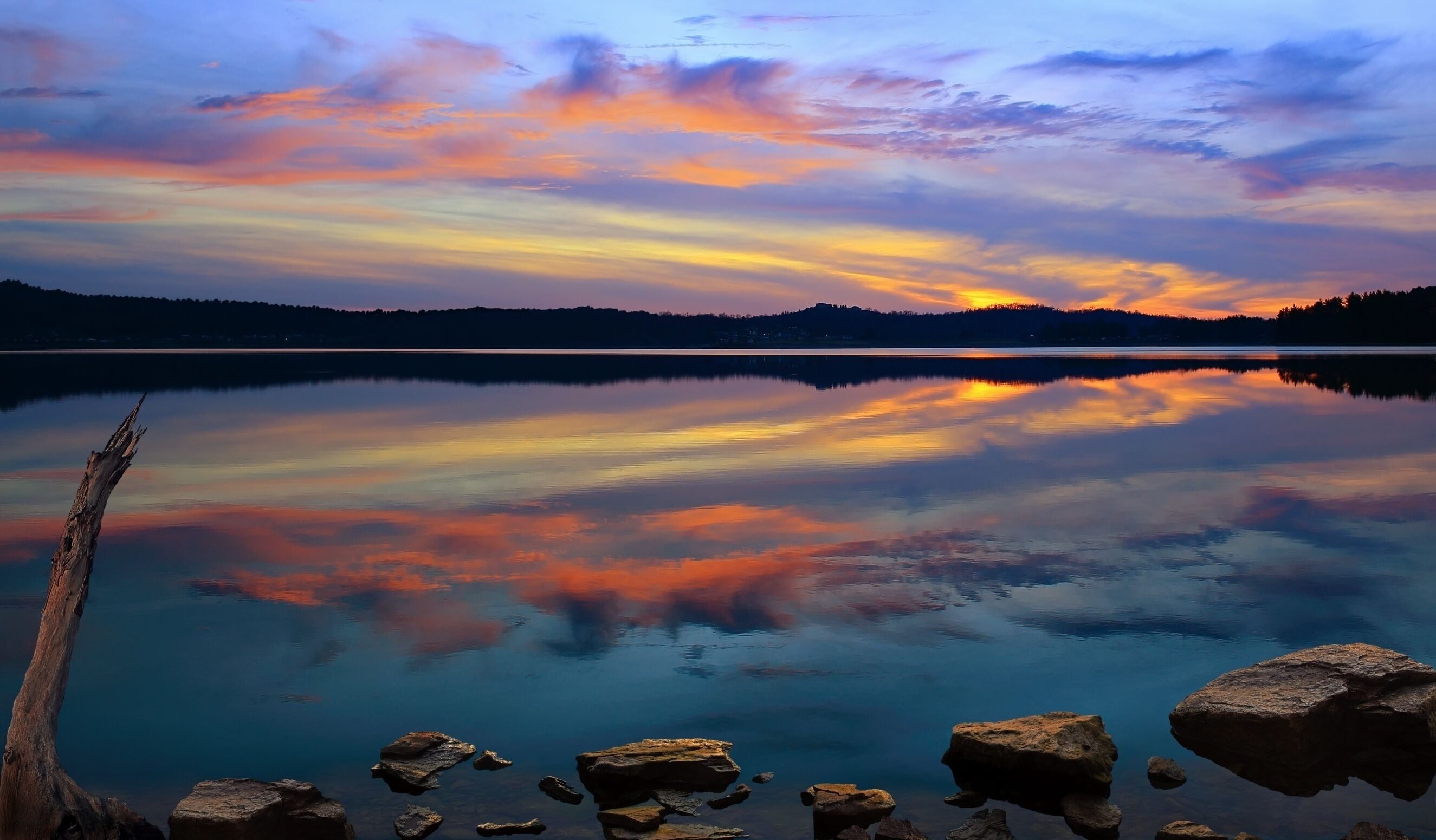 A calm lake with a sunset reflection, surrounded by rocks and a forested hill.