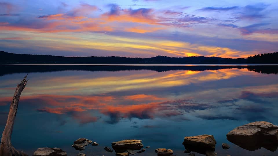 A calm lake with a sunset reflection, surrounded by rocks and a forested hill.