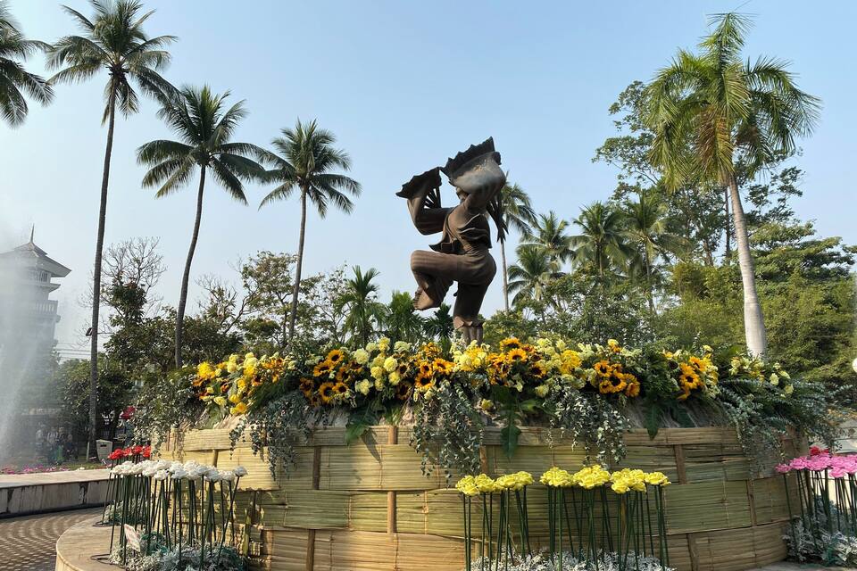 Uma estátua de uma criança com um balde, rodeada por flores vibrantes e palmeiras.