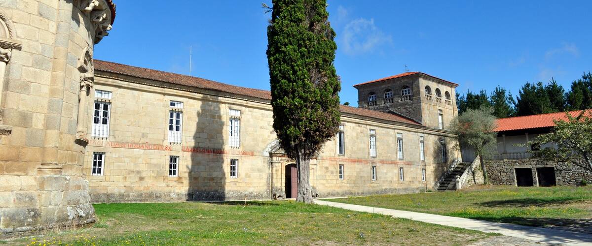 A historic stone building with a tall tree in front, a clear blue sky, and a red-roofed tower.