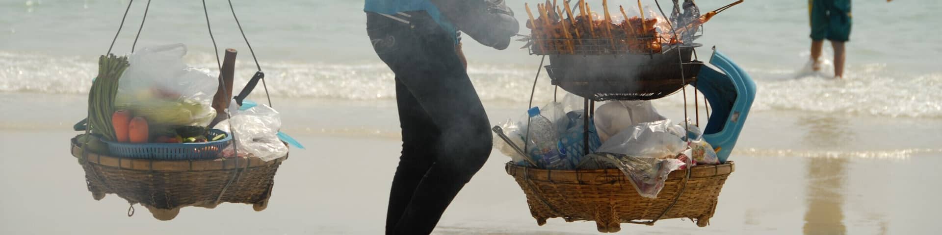 A woman carrying two baskets on a beach with a boat and people in the background.