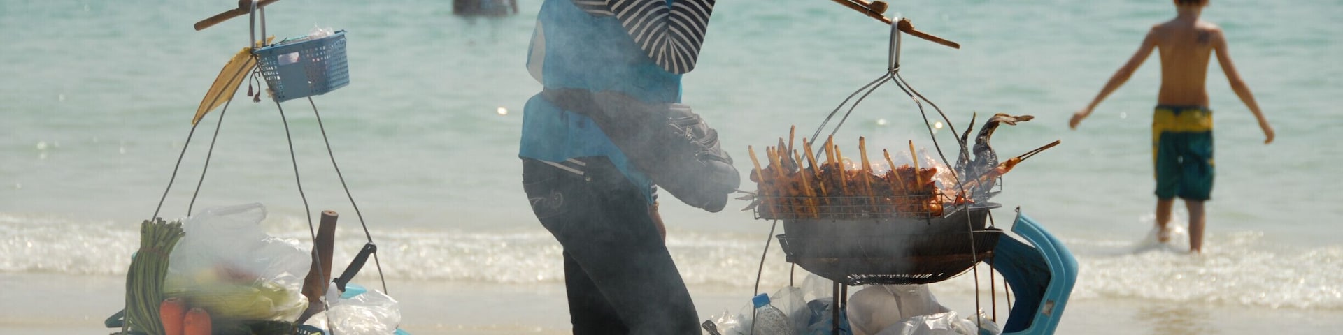 Una mujer lleva dos cestas en una playa, con un barco y gente de fondo.