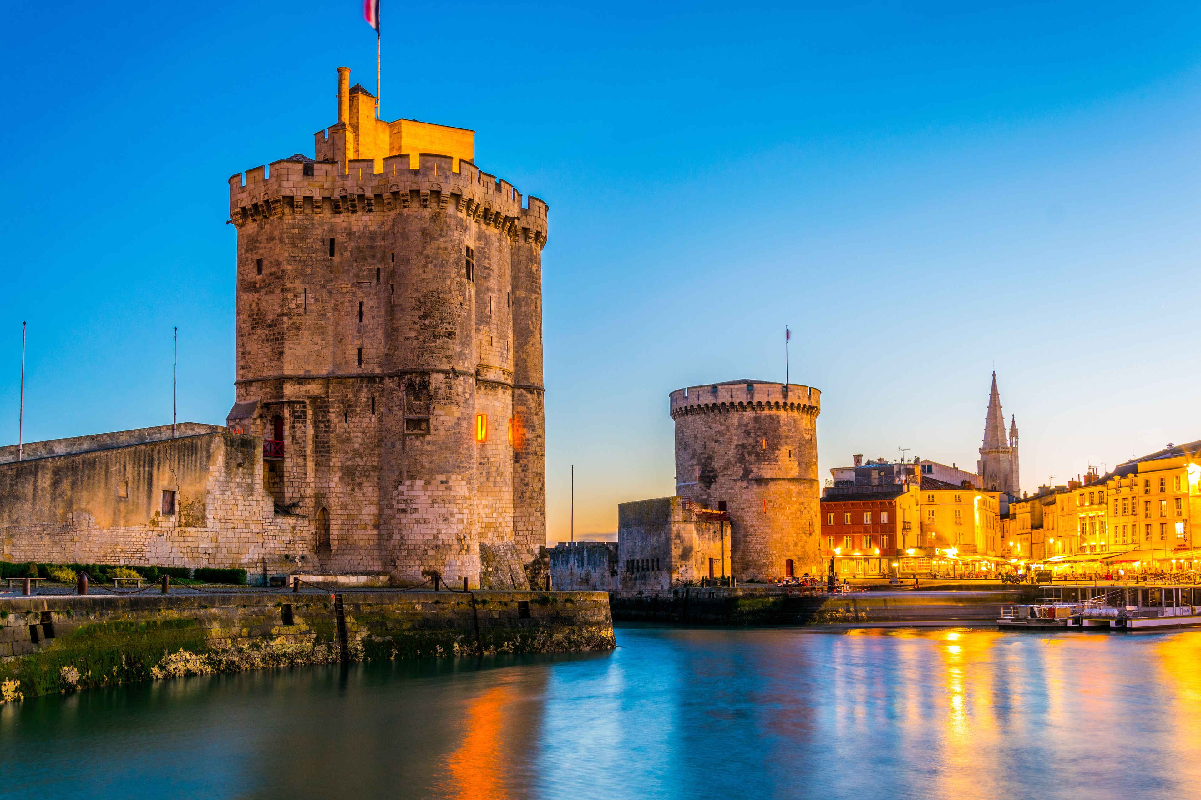 A historic castle with a flag, a tower, and a cityscape with a church in the background.