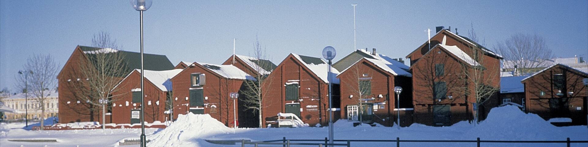 A snow-covered landscape with a metal fence, a snow pile, and several brick buildings with snow on their roofs.