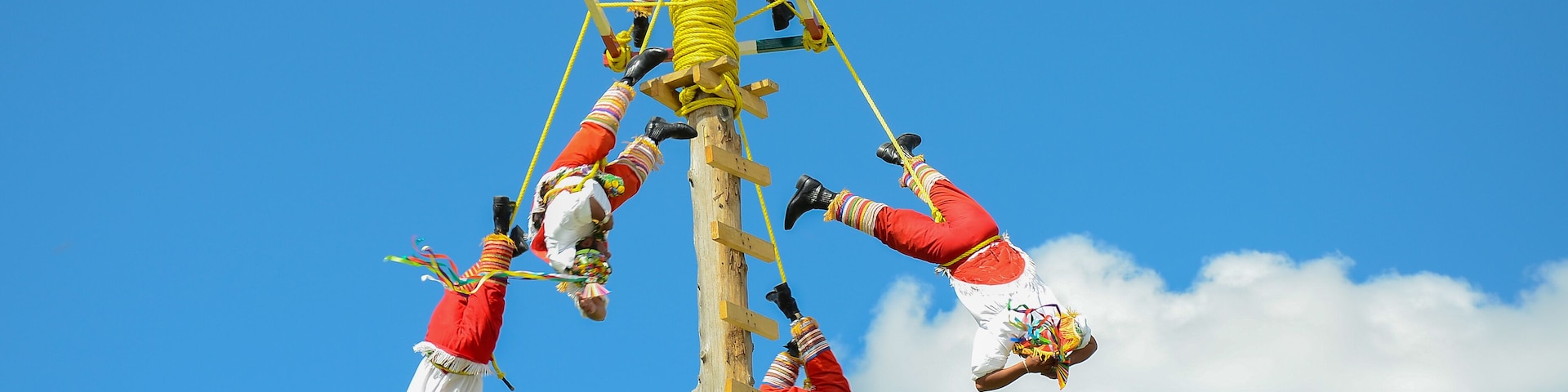 Un grupo de personas vestidas con atuendos tradicionales realizando una acrobacia aérea contra un cielo azul despejado.