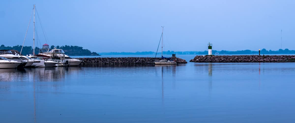 A calm harbor with several boats docked, a lighthouse, and a rocky breakwater.