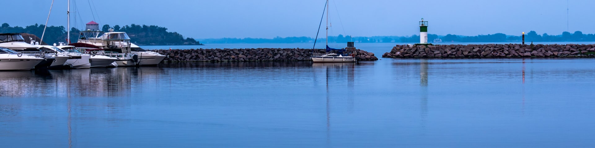 A calm harbor with several boats docked, a lighthouse, and a rocky breakwater.