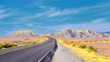 A winding road through a desert landscape with yellowish vegetation and rocky mountains in the background.