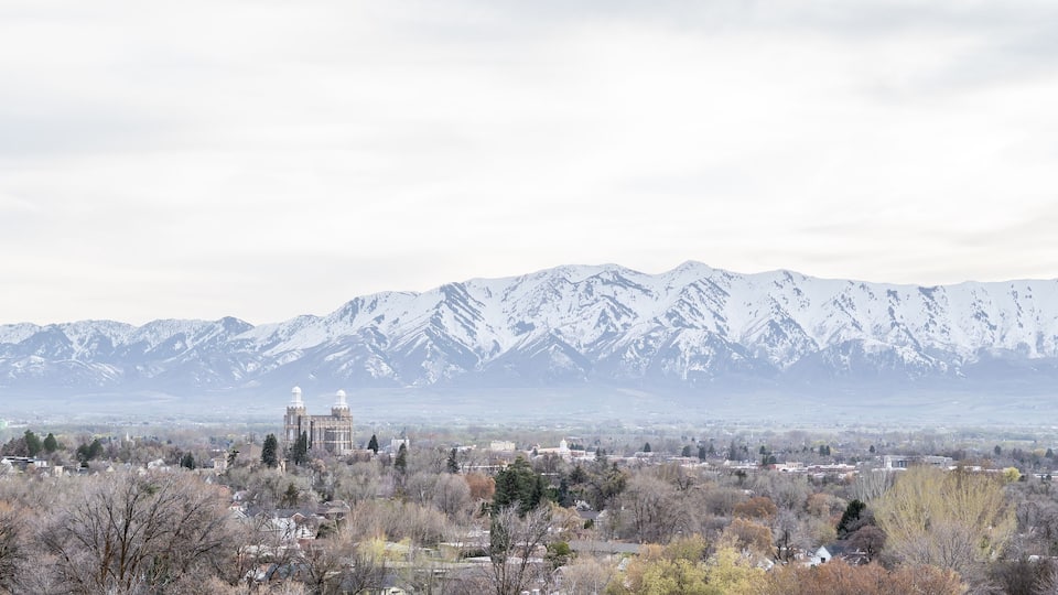 A town with a prominent building featuring two domes, surrounded by snow-capped mountains and autumn-colored trees.