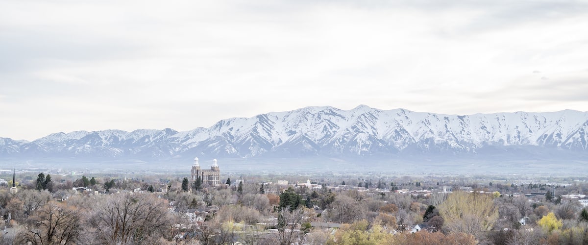 A town with a prominent building featuring two domes, surrounded by snow-capped mountains and autumn-colored trees.