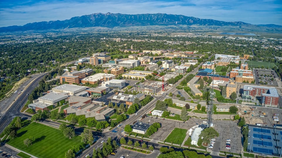 An aerial view of a city with a mix of modern and older buildings, surrounded by green spaces and a mountain range in the background.