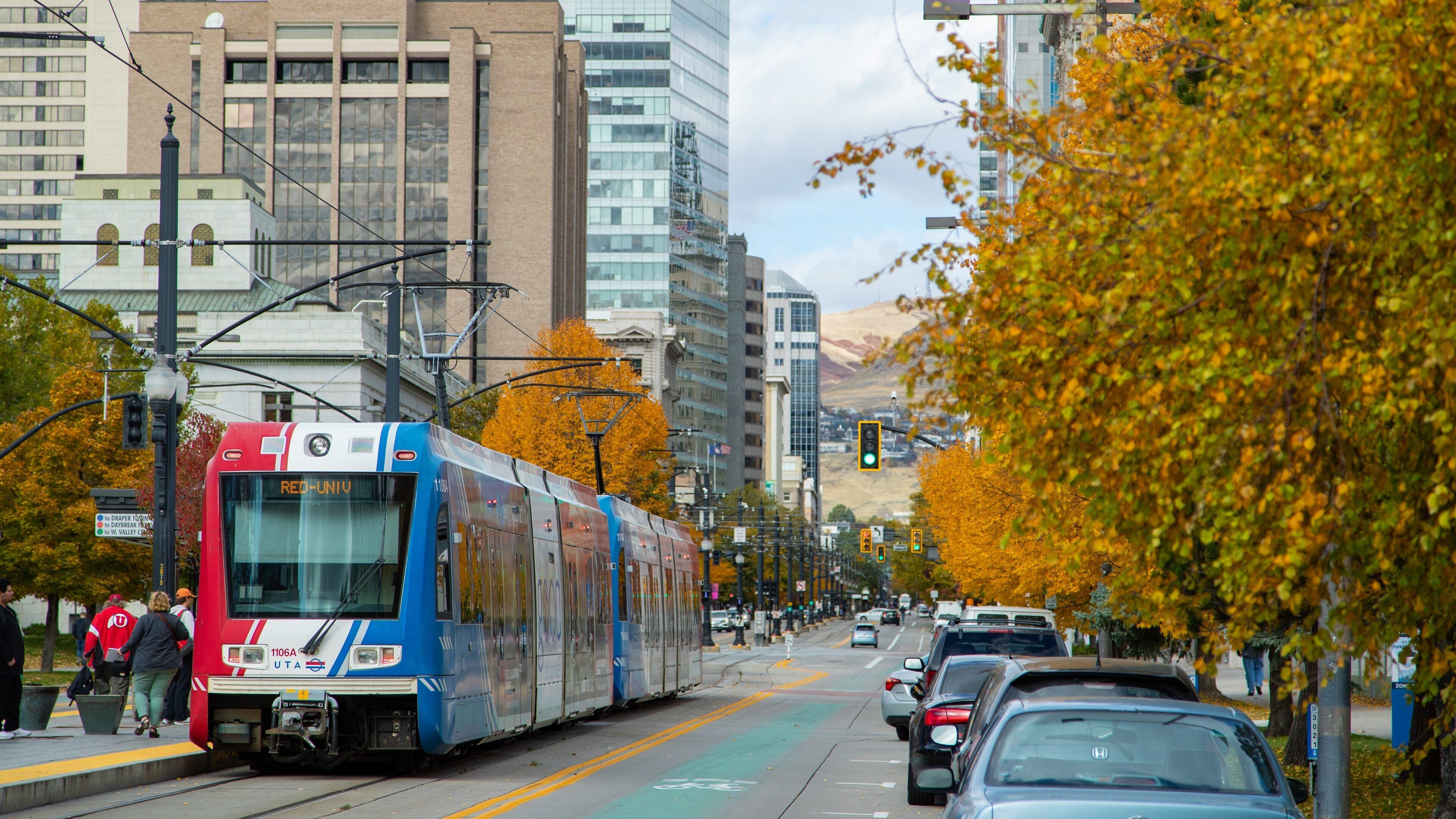 A red and blue tram on a city street with autumn trees and tall buildings in the background.
